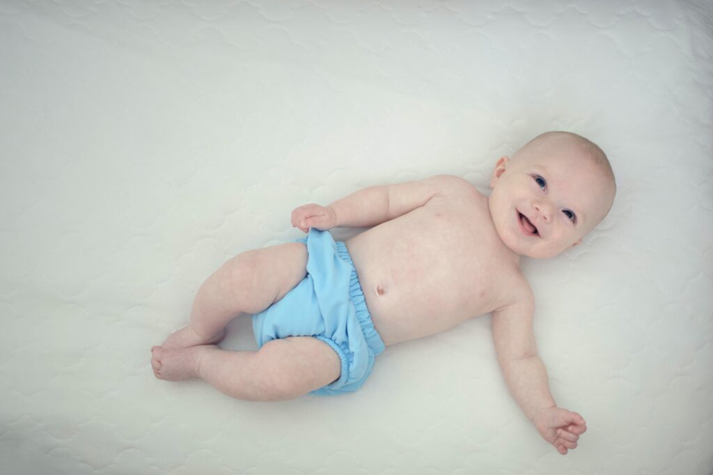A baby wearing a blue diaper cover smiling while laying on a white background for a post about Pampers Baby Dry vs Swaddlers